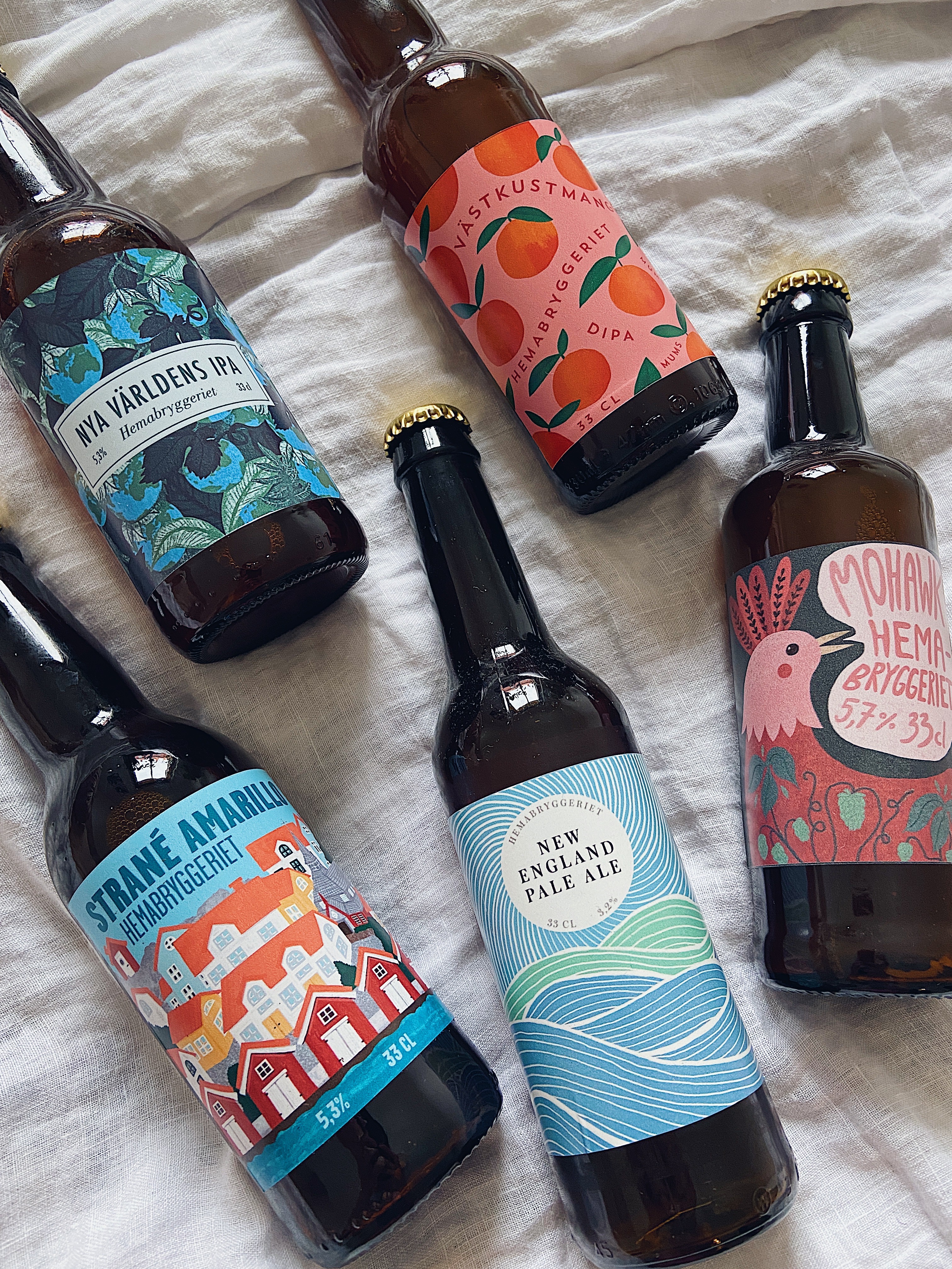 Fem olika ölflaskor med varierande etiketter, t.ex. med mango, jordglober, Hunnebostrand, vågor och en tupp.