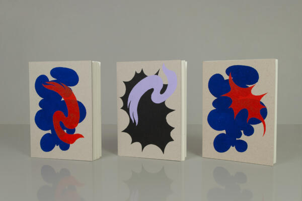 Tre handbunda och handtryckta skissböcker står på en blankgrå yta. Abstrakta motiv på omslagen i rött, blått och syrenlila.