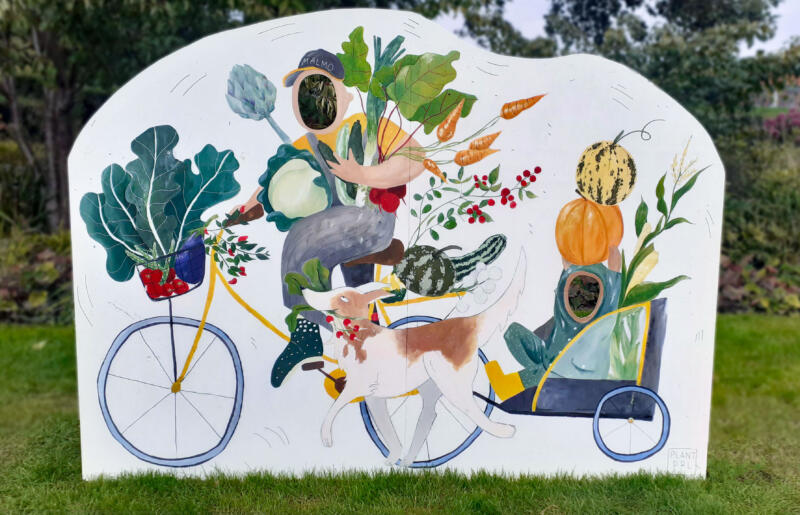 Offentlig målning föreställandes en person som cyklar. Personen har famnen och cykeln full av grönsaker. Ett barn i en cykelkärra håller i pumpor och en hund springer bredvid bärandes på rädisor.