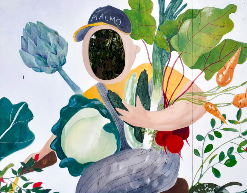Offentlig målning föreställandes en person som håller zucchini, purjolök, rödbetor och mer grönsaker i famnen