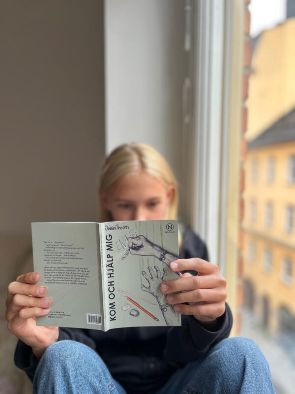 Flicka som sitter i fönster och läser boken "Kom och hjälp mig" av Johan Theorin. Boken är ljust grön och har en illustration av två händer på sig.