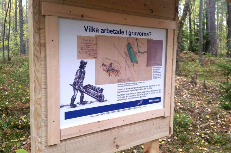 En av träskyltarna om Hållets gruvor i en skog i Nyköpings kommun. På skylten står det bland annat "Vilka arbetade i gruvorna?"