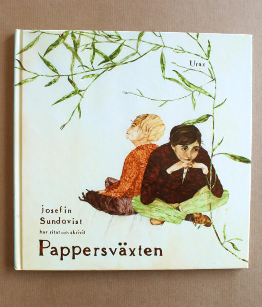 Foto av boken Pappersväxten. Handtextad titel och illustrerat bokomslag. Två barn med ryggarna mot varandra och en växt som klättrar över deras huvuden.