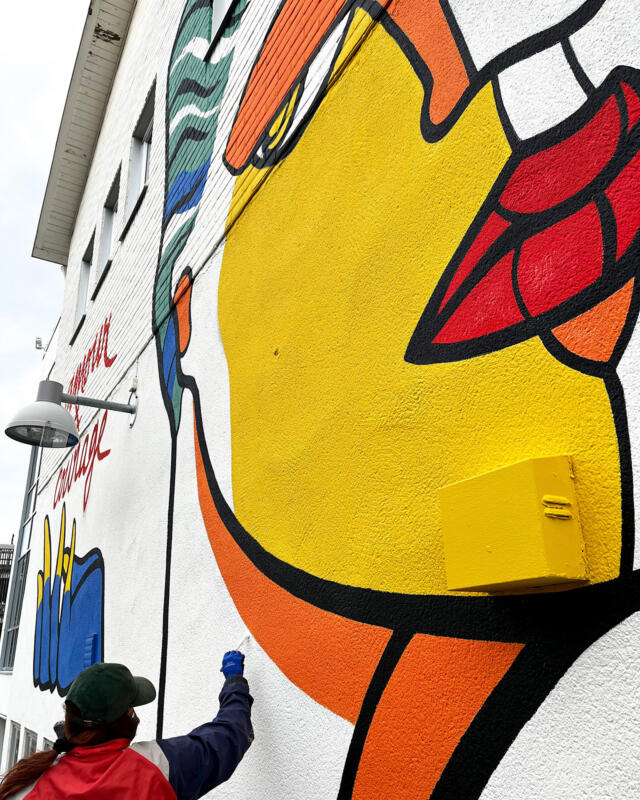 Närbild på muralmålning under arbete. Ett ansikte, en mun i flera färger samt en människa som målar den  vita bakgrunden. 