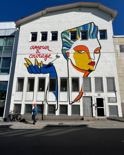 Vit byggnad med en muralmålning som representerar ett ansikte i olika färger samt en hand som håller upp texten "Amour & Courage". En människa går förbi byggnaden. 