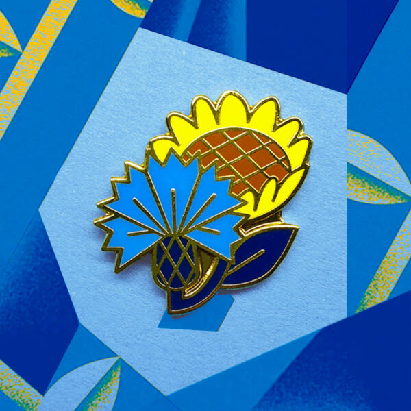 Emaljnålen som föreställer en blåklint och en solros på en blå bakgrund.