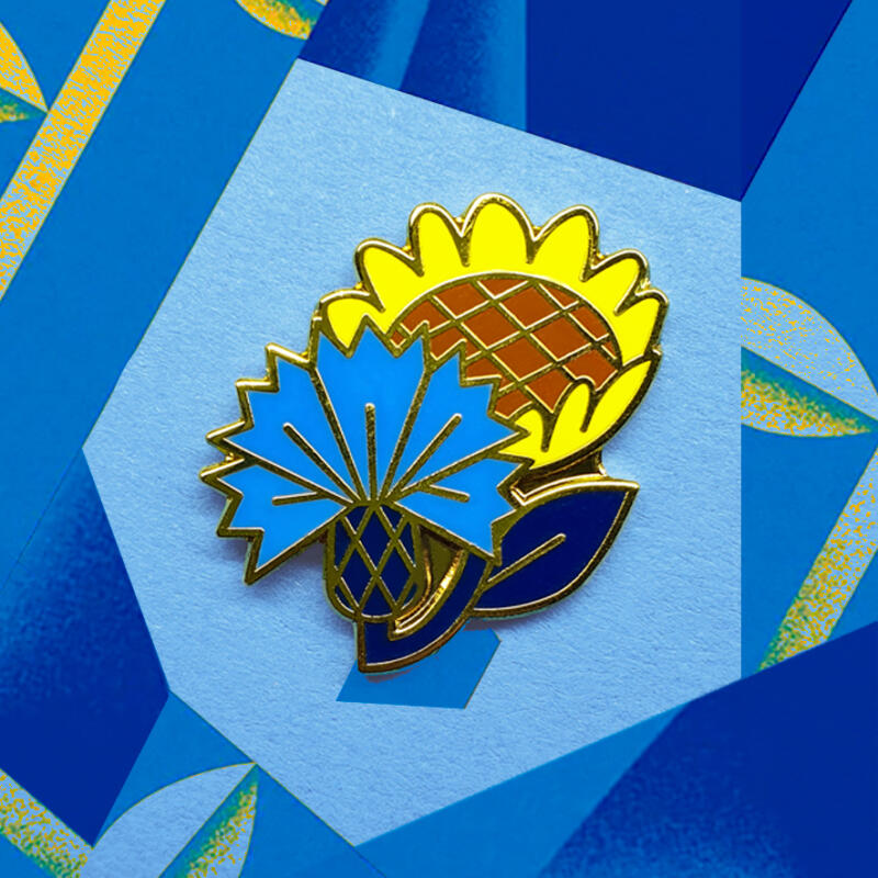 Emaljnålen som föreställer en blåklint och en solros på en blå bakgrund.