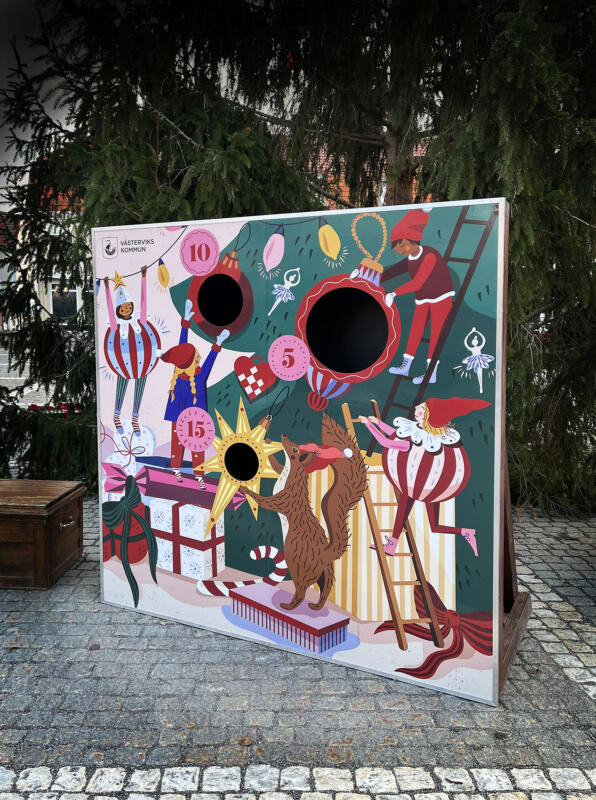 Foto av illustrationen till julspelen. Illusrtationen föreställer barn och djur som klär granen med pynt, julkulor och ljusslingor. I bilden ligger även paket och polkagrisar med.