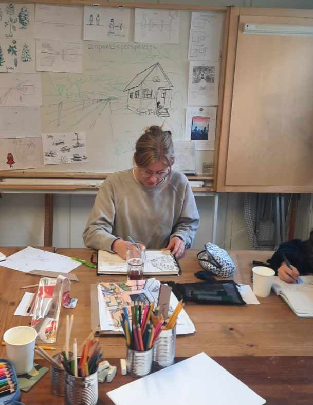 Elin Alskog håller en workshop där eleverna får rita manga, karaktärer, perspektiv och serier. De sitter vid ett bord där det finns teckningar, papper och pennor. I bakgrunden syns en whiteboard med exempelbilder och förklaringar av enpunktsperspektiv. 