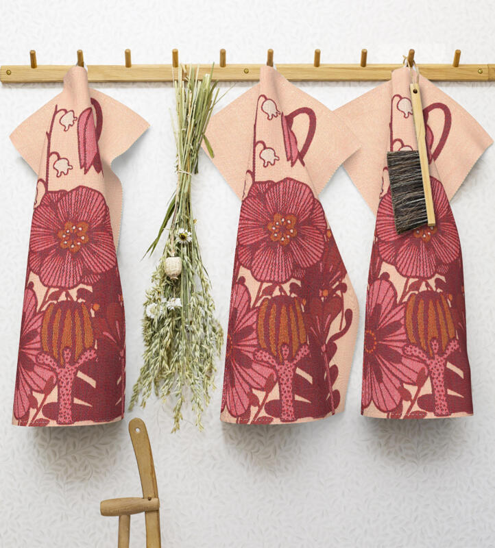 Bild på tre vävda kökshanddukar hängande på en knopplist tillsammans med en hängande bukett och borste. Vit tapet. Handdukarnas motiv är fantasifulla blommor i rosa, vinrött och brungult på en bakgrund som är ljusrosa/beige. Bakom blommorna sträcker några liljekonvaljer sig upp.