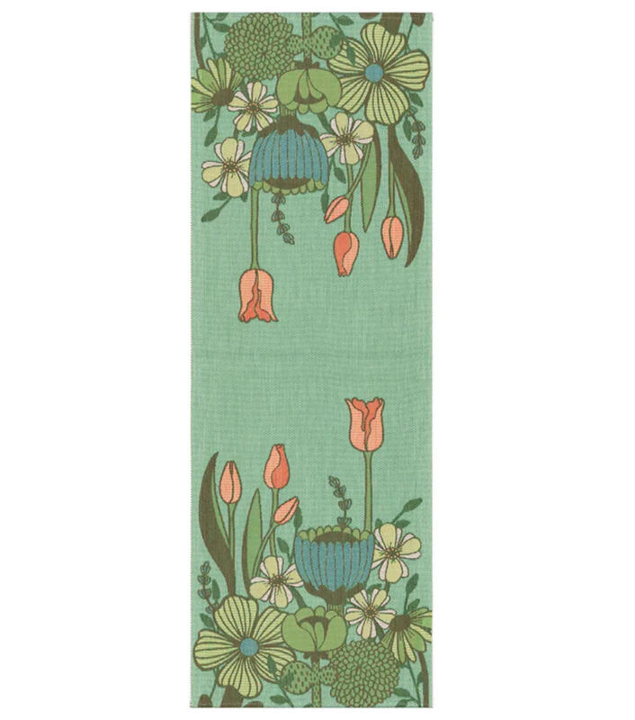 Vävd bordslöpare i mjuka gröna nyanser och olika fantasifulla blommor. Fyra tulpaner i rosa sträcker sig upp bakom de andra blommorna. Motivet är speglat i varsin ände av duken med bara bakgrundsfärg i mitten.