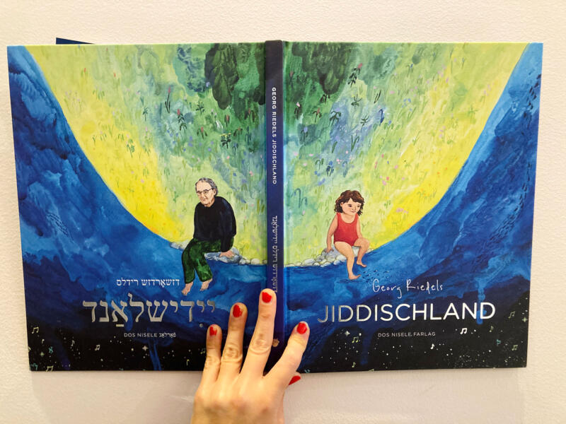 Omslag till Georg Riedels bok ”Jiddischland”, en vändbar bok som kan läsas på jiddisch eller svenska. Framsidan visar en flicka, baksidan Georg Riedel. Boken gestaltar livets flod och kombinerar berättande med konstnärlig illustration för att förmedla livets gång, kulturarv och personliga upplevelser på ett visuellt tilltalande sätt.