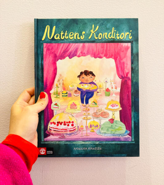 Bokomslag barnbok, bilderbok för barn 3-6 år. Handgjord, akvarell och collage föreställande ett konditori med ett fönster fullt av bakelser och en pojke som äter kakor, tårtor, jordgubbar, bakelser