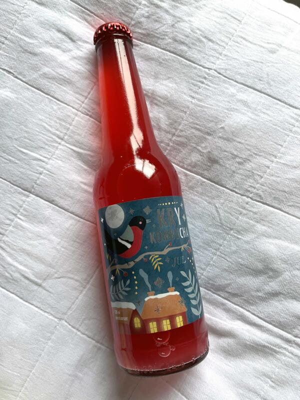 En julig etikett på en kombuchaflaska från Norge med röd dryck i. Juletiketten har motiv av en domherre, hus med lysande fönster, rönnbär, måne och snöflingor.