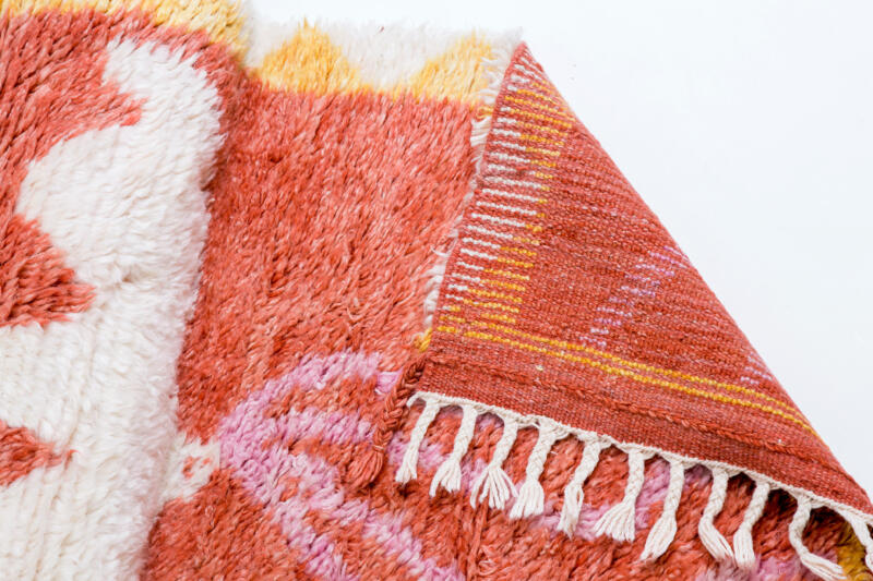 Närbild, produktbild, av berbermatta med fokus på ena hörnet som är uppvikt och visar det handknutna hantverket bakom. Flätade fransar. Mattan är korallrosa med vit och gul bård i trianglar, vit fågel med rosa band i näbben.