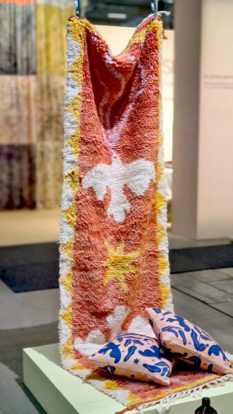 Handknuten berbermatta, lång gångmatta med flätade fransar i kortändan. Upphängd på display för hantverksutställning. Mattan har vita och varmt gula trianglar längs långsidorna, innanför trianglarna är bakgrunden korallrosa med en gul stjärna i mitten, två vita fåglar på varsin sida om den, hållande rosa band i näbbarna.