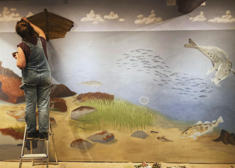 En kvinna målar en träeka (båt) som vilar på vattenytan i en jättestor väggmålning, som visar havet så som det så ut på förr, i lekutställningen Gammeldagstiden i Österlens museum, Simrishamn. Under ytan kryllar det av ålar, sillar och torskar. Här finns även gråsäl och lax.