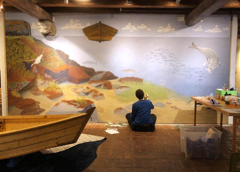 En kvinna sitter och målar detaljer på en jättestor väggmålning, som visar havet så som det så ut på förr, i lekutställningen Gammeldagstiden i Österlens museum, Simrishamn. Det kryllar av ålar, sillar och torskar. Här finns även gråsäl och lax.