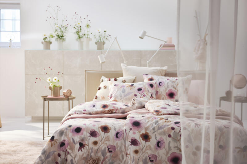 A bed made up with floral bed linen in a light and romantic setting. There are flowers in vases and a sheer curtain.
