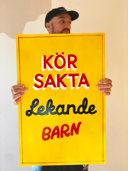 Man med keps håller en handmålad skylt med texten "Kör sakta, lekande barn". Skylten är gul med text i rött, vitt och svart.