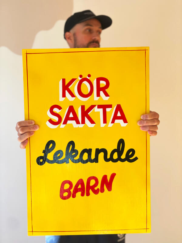 Man med keps håller en handmålad skylt med texten "Kör sakta, lekande barn". Skylten är gul med text i rött, vitt och svart.