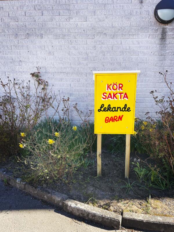En handmålad skylt med texten "Kör sakta, lekande barn" står i en rabatt framför en byggnad. Skylten är gul med text i rött, vitt och svart.