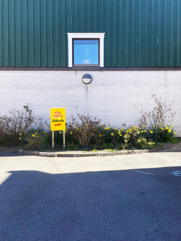 En handmålad skylt med texten "Kör sakta, lekande barn" står i en rabatt framför en byggnad. Skylten är gul med text i rött, vitt och svart.
