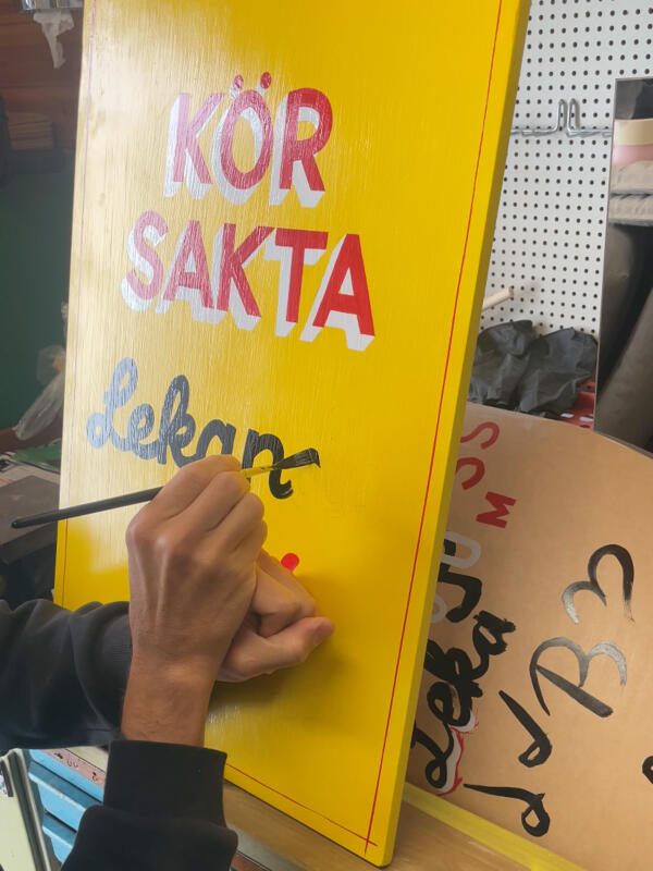 Processvideo av hur en handmålad skylt med texten "Kör sakta, lekande barn" skapas. Skylten är gul med text i rött, vitt och svart. Ett par händer som håller en pensel syns.
