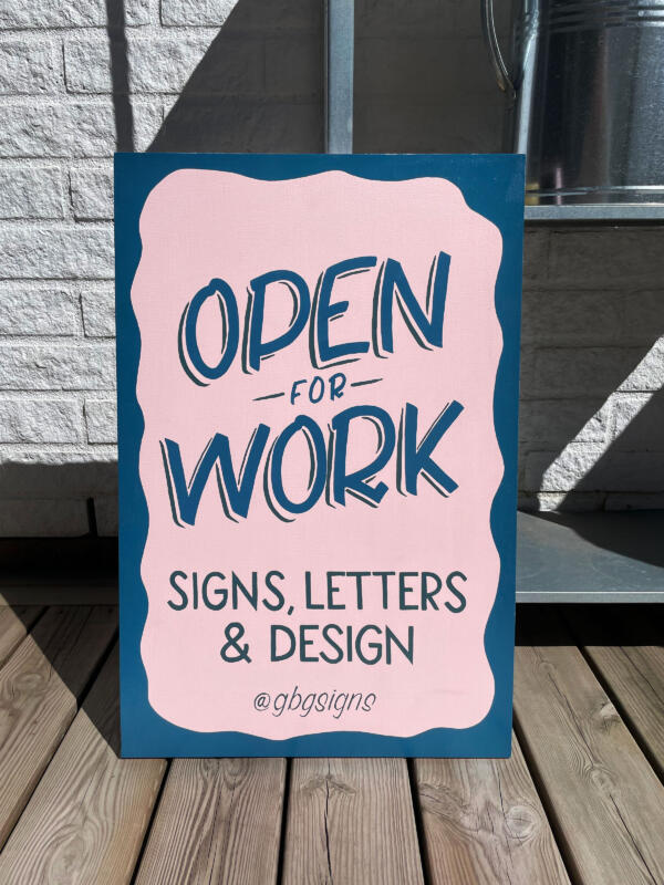 Handmålad skylt med text som står på en trätrall utomhus, lutad mot en hylla framför en tegelvägg. Skylten är rosa med blå bokstäver och en målad ram. Texten säger "Open for Work" och "Signs, Letters & Design" samt instagramnamnet @gbgsigns