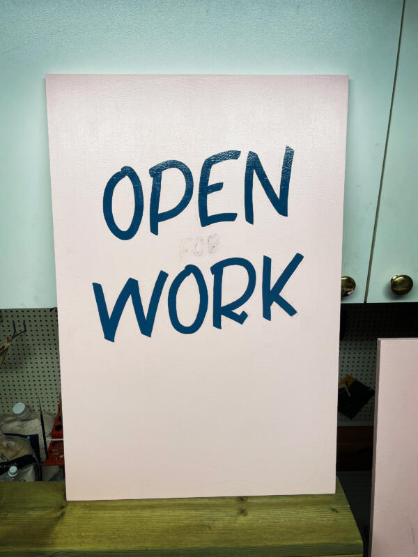 Handmålad skylt med text som står på en bänk i verkstadsmiljö. Skylten är rosa med blå bokstäver. Texten säger "Open for Work"
