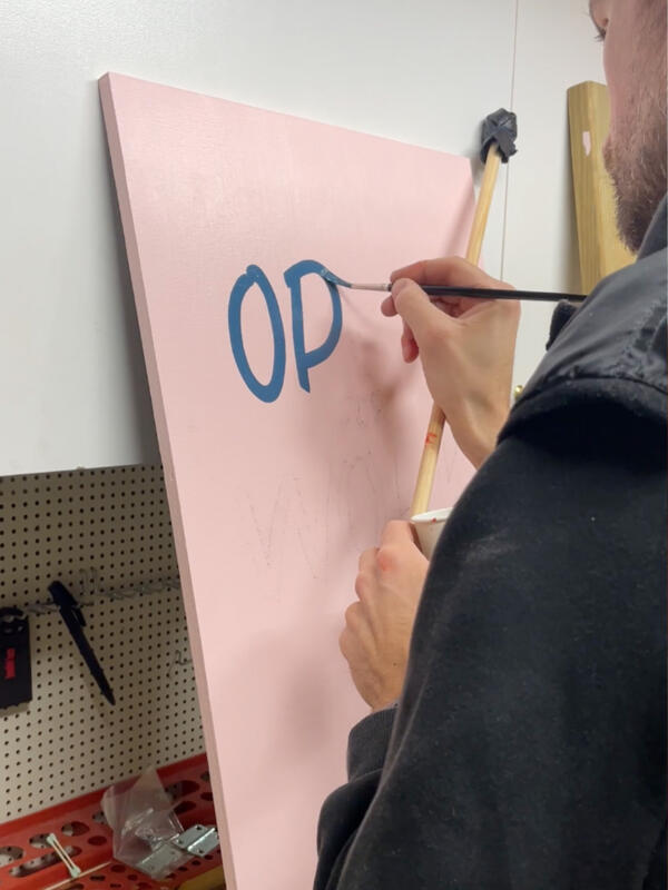 Handmålad rosa skylt med text som står på en bänk i verkstadsmiljö. En man målar blå bokstäver med pensel. Han håller även i en "mahl stick"
