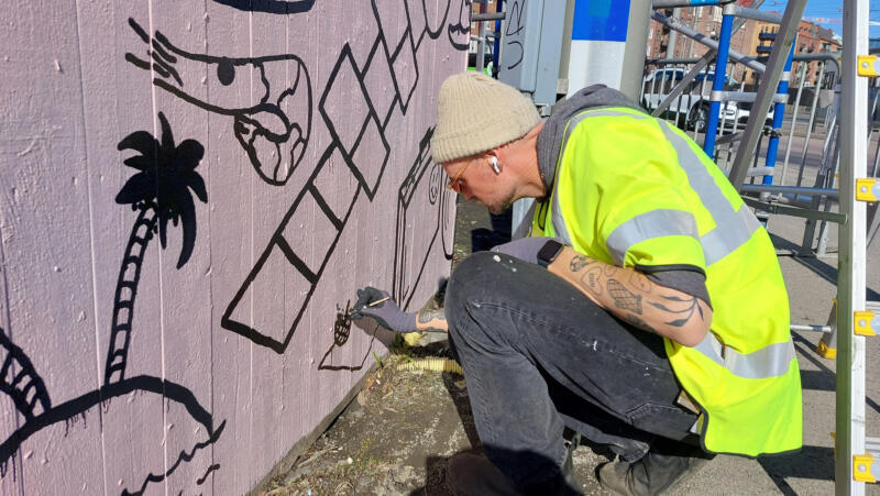 Väggmålning / muralmålning på del av ett byggplank vid Korsvägen i Göteborg. Rosa bakgrund med många svarta illustrationer, eller doodles, i olika storlekar. Bild av processen, man som målar på väggen.