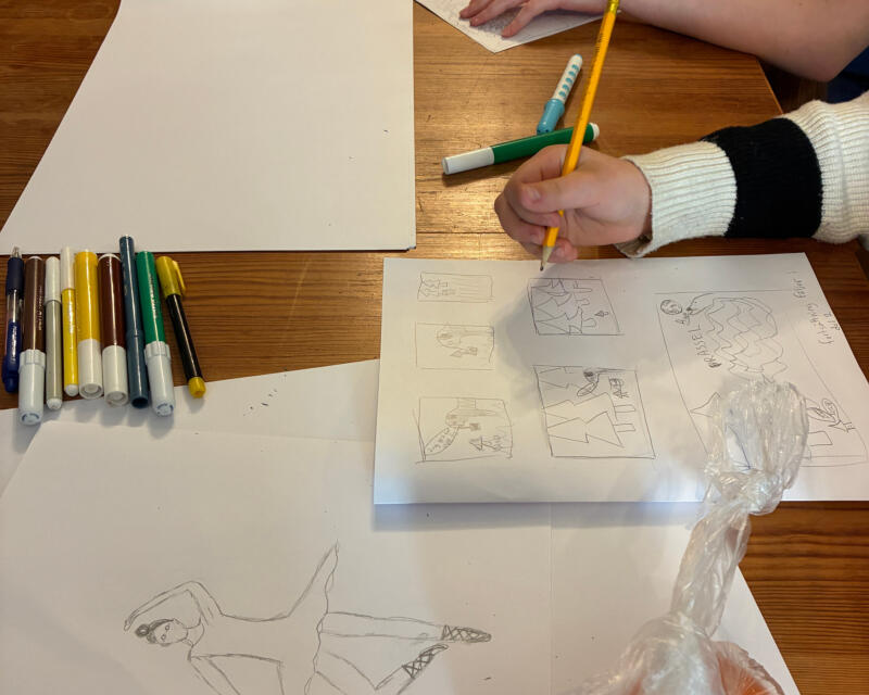Ett bord med papper och pennor. En teckning av en ballerina och ett papper med en ritad serie på. En deltagare håller på och ritar på serien.
