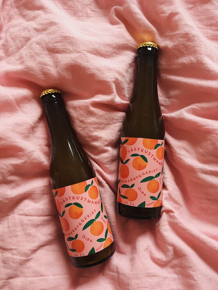 Två ölflaskor med etiketter av mango mot en rosa bakgrund, texten säger "Västkustmango" och "Dipa" och "mums" bland annat.