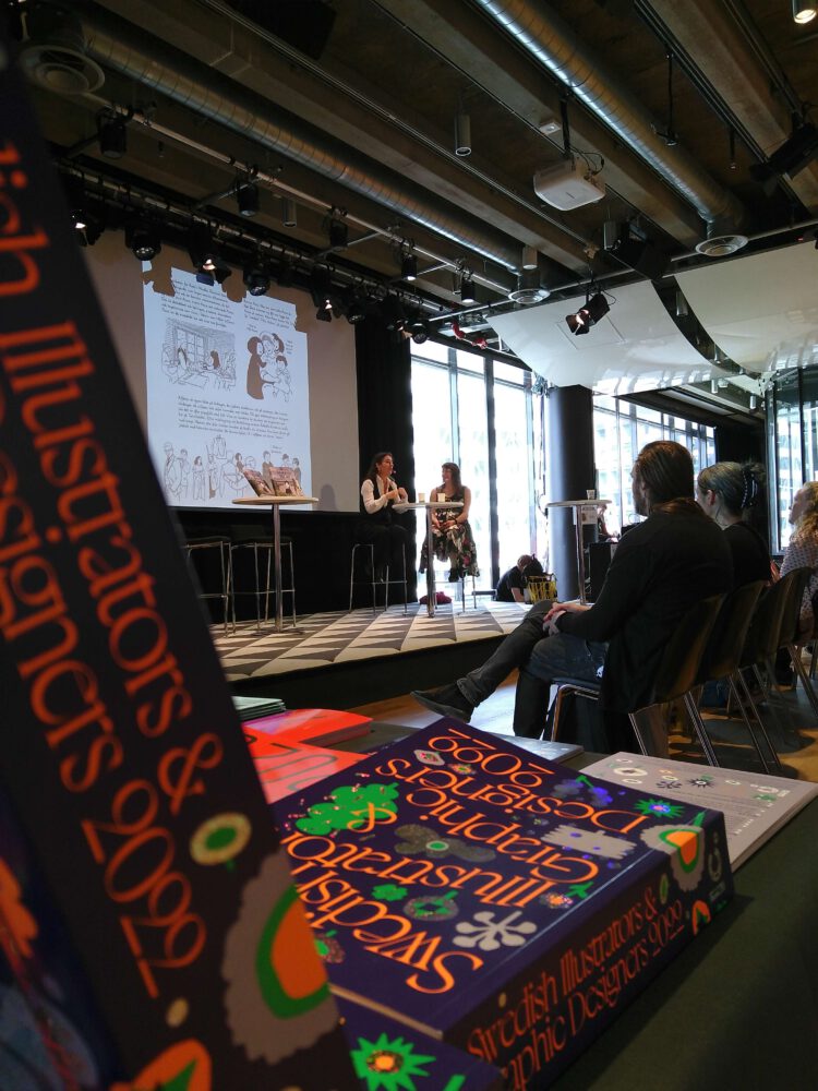 Bilden visar Illustratörcentrums katalog "Swedish Illustrators & Graphic Designers" i förgrunden. I bakgrunden ser vi en scen med två personer i samtal på stockholms internationella seriefestival 2022. 