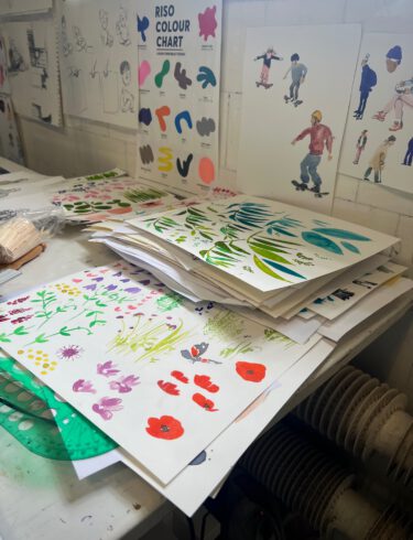 Bilden visar ett rum med färgglada illustrationer som ligger utspridda på ett vitt bord och upphängda på en vit vägg. Motiven är bland annat blommor, en person som åker skateboard.
