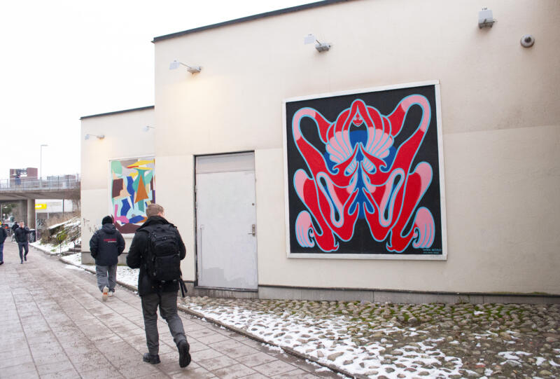 Människor går förbi en muralmålning i stor ram på en beige husfasad. Motiv i ramen föreställande en röd kvinnofigur med ljusblå konturer mot svart bakgrund.