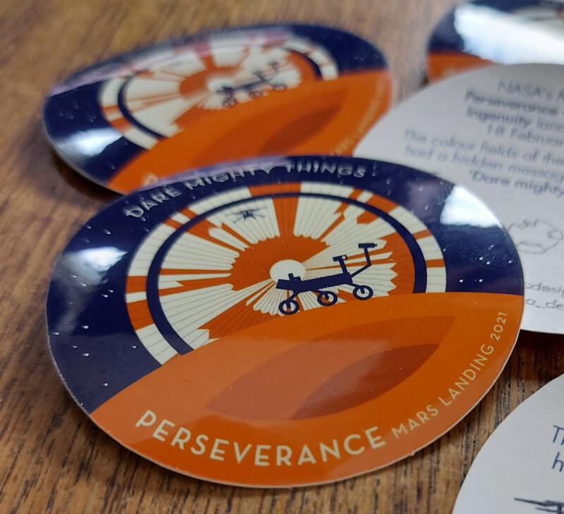 Photo of Perseverance stickers. One in the front is in focus and takes most of the space. To the right you see part of an upside down sticker with back paper print about the mission.