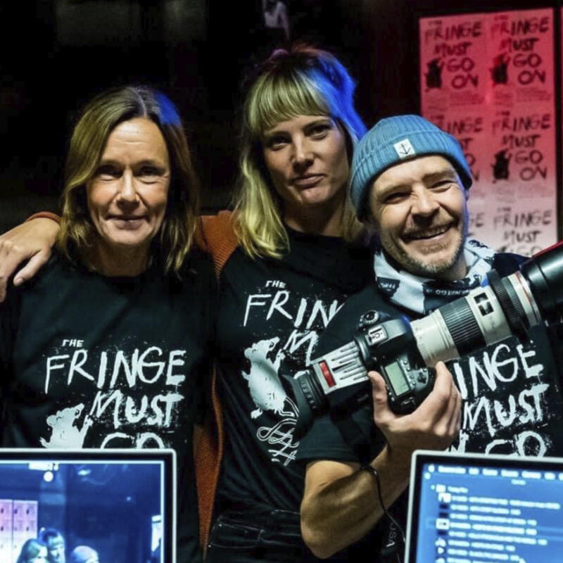 Tre personer med t-shirts där det står »The fringe must go on«. I bakgrunden syns affischer med samma budskap.