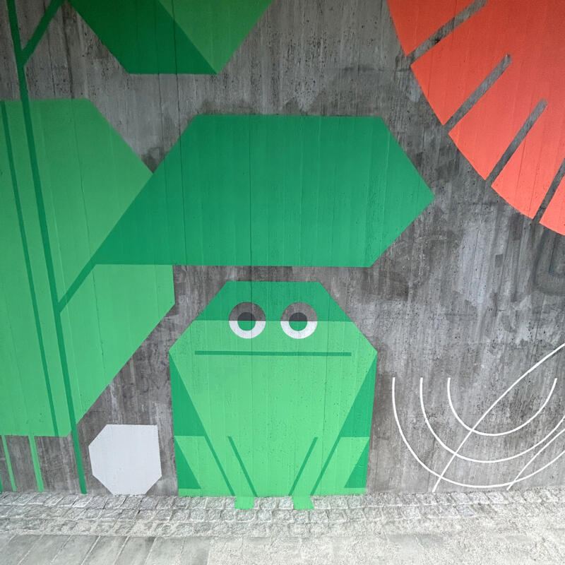 Muralmålning på grå betongvägg föreställande en grön groda.