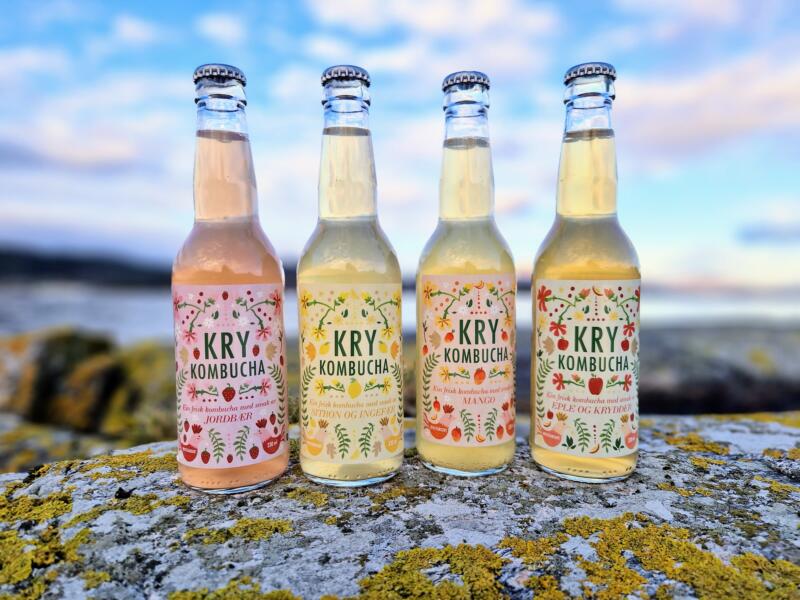 Fyra olika norska kombuchaflaskor med etiketter för olika smaker: jordgubb, citron och ingefära, mango, äpple och kryddor.