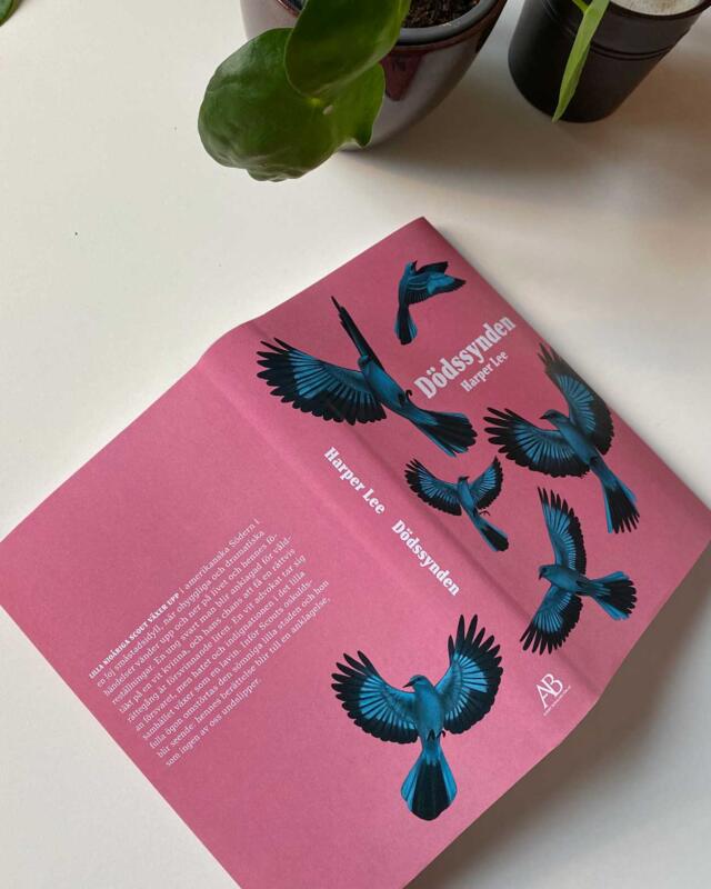 Bok ligger uppslagen på ett bord. Omslagsdesignen visar blå fåglar mot en rosa bakgrund.