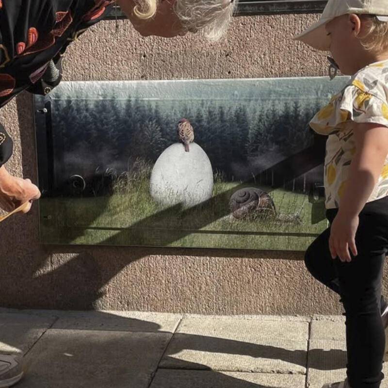 En kvinna och ett barn på varsin sida av det applicerade motivet Ägget. Ett fotocollage med en pilfink sittandes på ett stort ägg på en äng, omgiven av en mörk skog. Framför ägget kryper en snigel förbi.