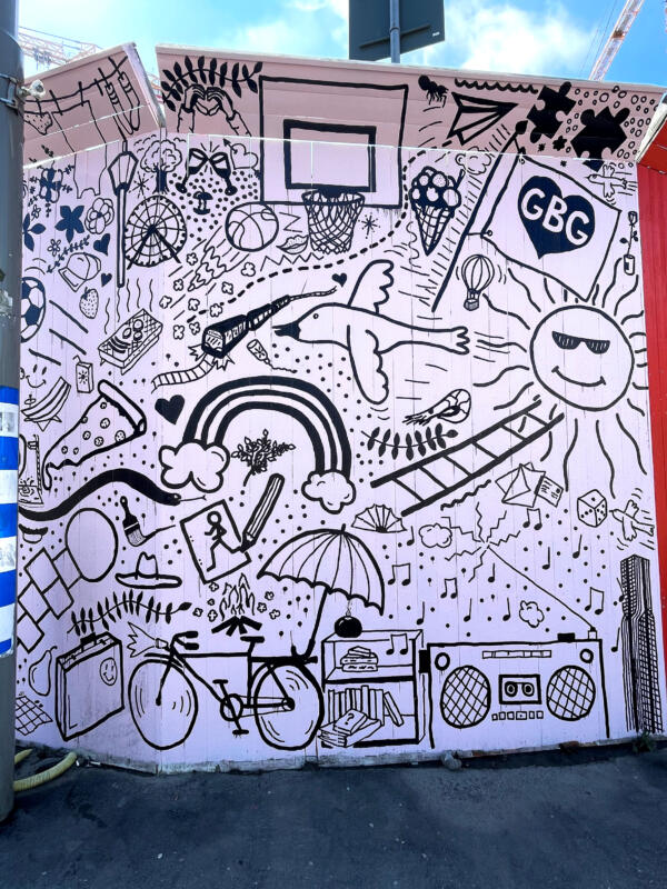 Väggmålning / muralmålning på del av ett byggplank vid Korsvägen i Göteborg. Rosa bakgrund med många svarta illustrationer, eller doodles, i olika storlekar. 