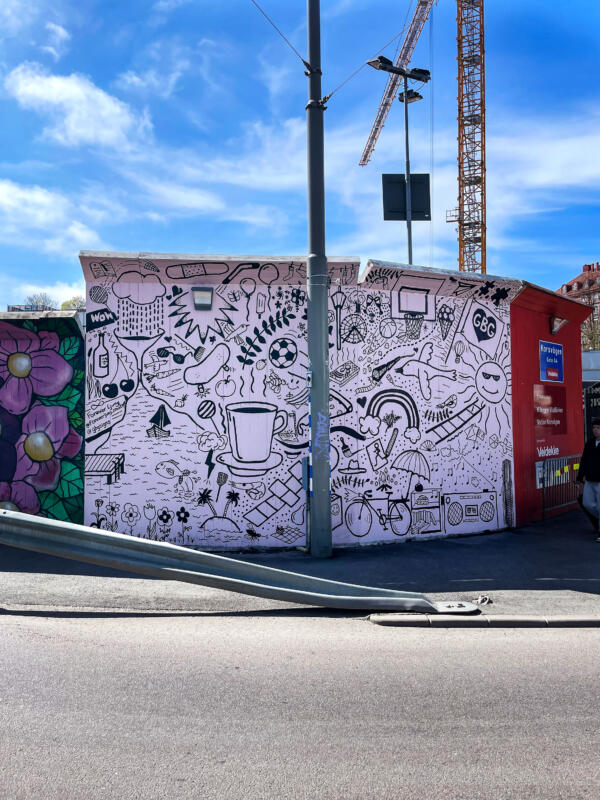 Väggmålning / muralmålning på del av ett byggplank vid Korsvägen i Göteborg. Rosa bakgrund med många svarta illustrationer, eller doodles, i olika storlekar. 