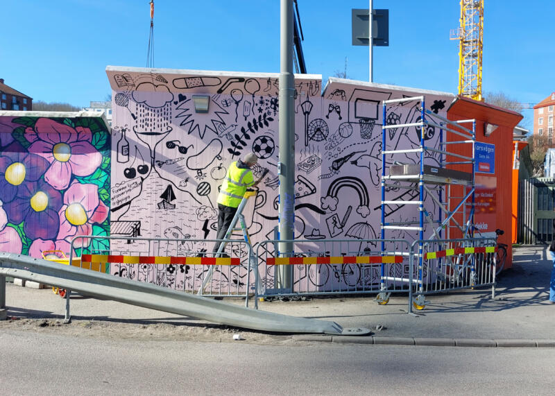 Person på stege målar en väggmålning på ett byggplank vid Korsvägen i Göteborg. Framför väggen står ett byggstaket och en byggställning.