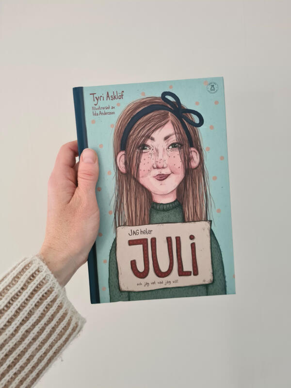 Omslagsbild på Juli till boken Jag heter Juli och jag vet vad jag vill. Juli tittar nyfiket och lite busigt på dig och presenterar sig med en skylt där bokens titel står.