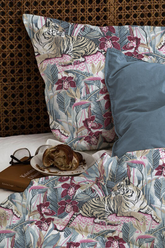 Interiörbild av uppbäddad säng med ett påslakan föreställande en vilande tiger omgiven av orkidéer, paradisblomma, och palmblad. Livliga färger i denimblått, vinrött, rosa och beige.