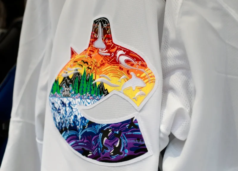 Photography of a close-up of a white hockey jersey with the Vancouver Canucks crest on it filled with a bright & colourful pride design depicting a landscape. 
