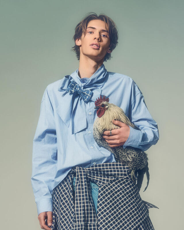 Boy in light blue shirt with a bow collar, checked shirt tied around waist and a rooster under his arm.